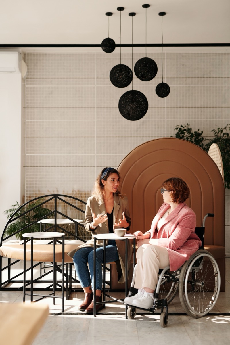 Two friends sitting together at a cafe table, one of them using a wheelchair.