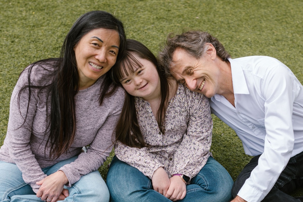 A family of three sitting together on the grass, leaning in and smiling.