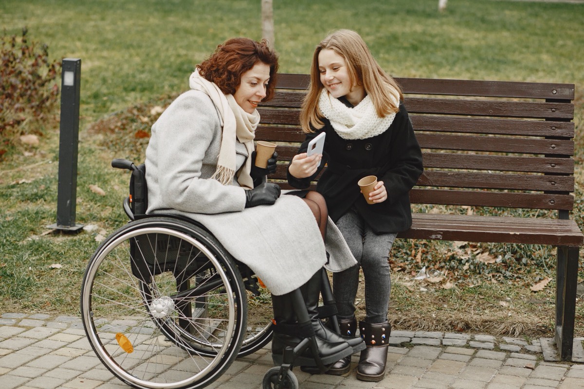 Two women talking together on a park bench outdoors, one of them using a wheelchair.
